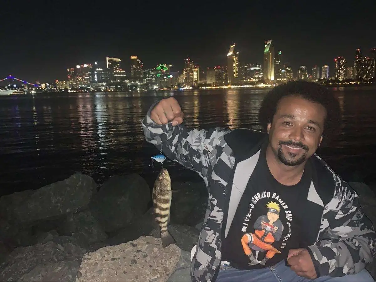 Crvne posing with a spotted bay bass he just caught at Bayview Park with the city of San Diego in the background, lit up against the night sky.